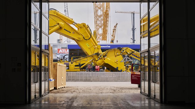 Eine große, gelbe Baggerschaufel steht hinter einem Eingangsbereich, umgeben von Kranen und Holzkisten.
