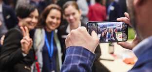 Ein Mann im karierten Anzug fotografiert mit einem Smartphone eine Gruppe von drei Frauen, die an einem Messestand posieren.