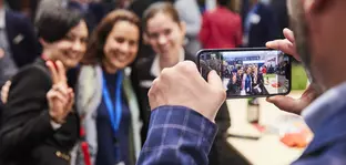 Ein Mann im karierten Anzug fotografiert mit einem Smartphone eine Gruppe von drei Frauen, die an einem Messestand posieren.