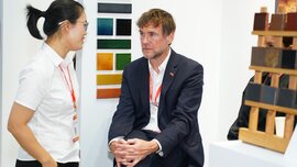 A man in a suit sits on a chair at a ceramitec exhibition stand while a woman looks at him and smiles.