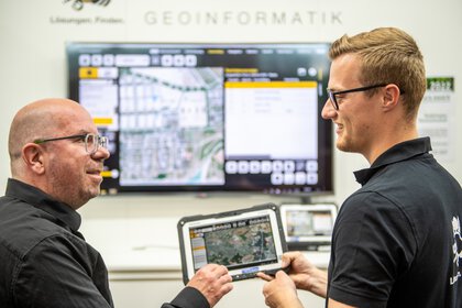 Zwei Männer diskutieren über Geoinformationsdaten, die auf einem Tablet und einem großen Bildschirm angezeigt werden.