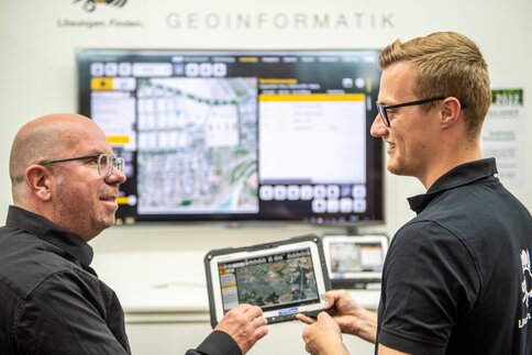 Zwei Männer diskutieren über Geoinformationsdaten, die auf einem Tablet und einem großen Bildschirm angezeigt werden.