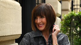 A smiling woman with shoulder-length brown hair is wearing a black denim jacket and black trousers and is standing in front of a yellow-brick building.