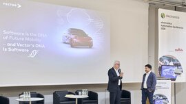 Two men are speaking on stage at a conference, while a slide about automotive software is shown behind them.