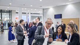 People networking at a conference stand around tables and chat in a modern, bright room.