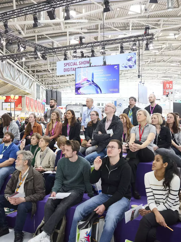 Auf der analytica sitzen und stehen viele Personen in einer Halle und folgen einer Special Show