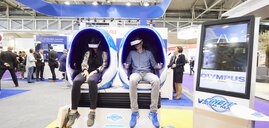 Two people sit in futuristic, egg-shaped chairs at a trade fair and use VR headsets.