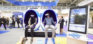 Zwei Personen sitzen in futuristischen, eiförmigen Stühlen auf einer Messe und nutzen VR-Headsets.