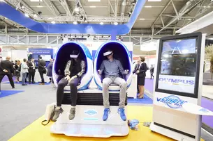Two people sit in futuristic, egg-shaped chairs at a trade fair and use VR headsets.