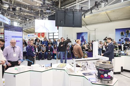 Trade fair visitors stand in front of a honeycomb-shaped table and look at exhibits