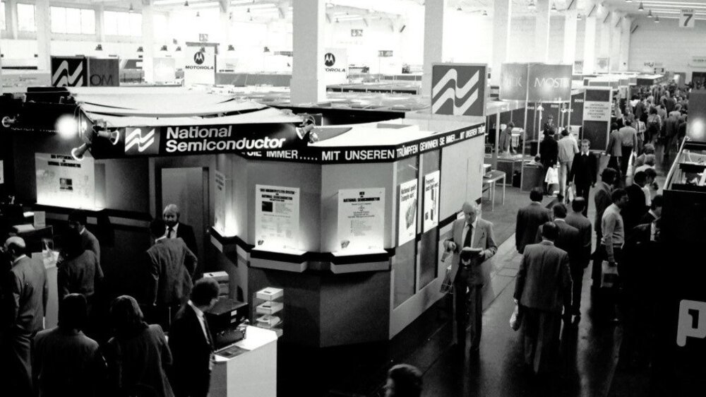 Black and white photo from an earlier productronica. People walk through a hall in which various exhibition stands are set up.