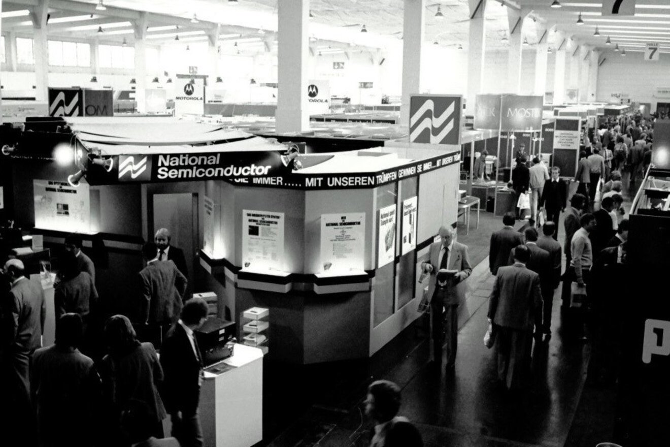 Schwarz-weißes Foto von einer früheren productronica. Menschen laufen durch eine Halle, in der diverse Messestände aufgebaut sind.