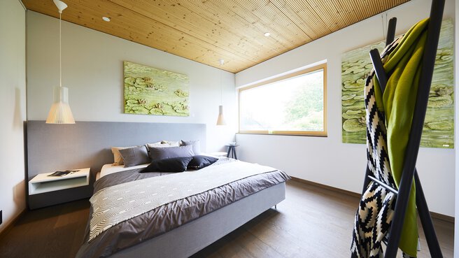 Modernes Schlafzimmer mit Holzdecke, großem Fenster, Doppelbett, grünen Kunstwerken und einer Leiter mit Decken.