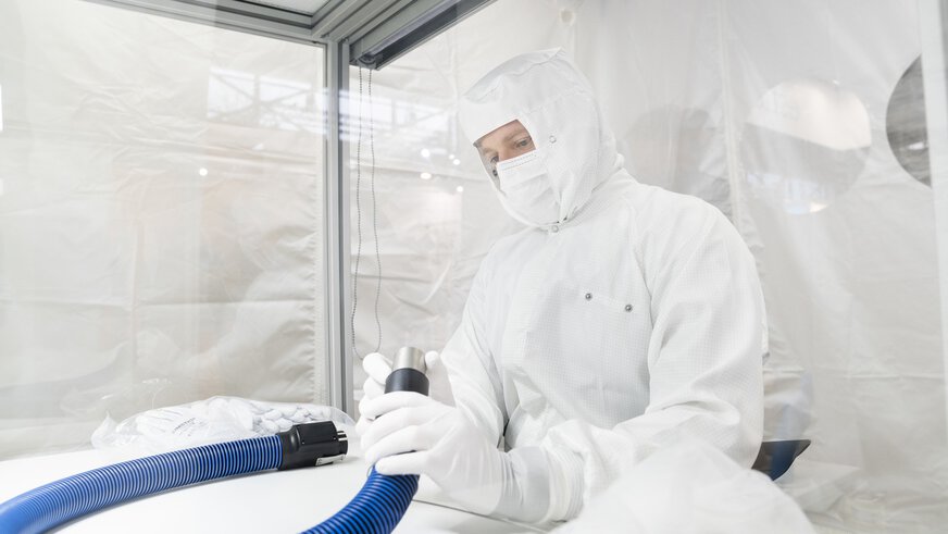 Eine Person in einem Schutzanzug, die in einer Reinraumumgebung sitzt und einen blauen Schlauch in der Hand hält.