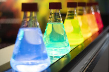 Fünf bunte, leuchtende Kolben in einer Reihe auf einer Laborbank. Jeder Kolben enthält eine fluoreszierende Flüssigkeit. Die Szene wirkt wissenschaftlich und experimentell.