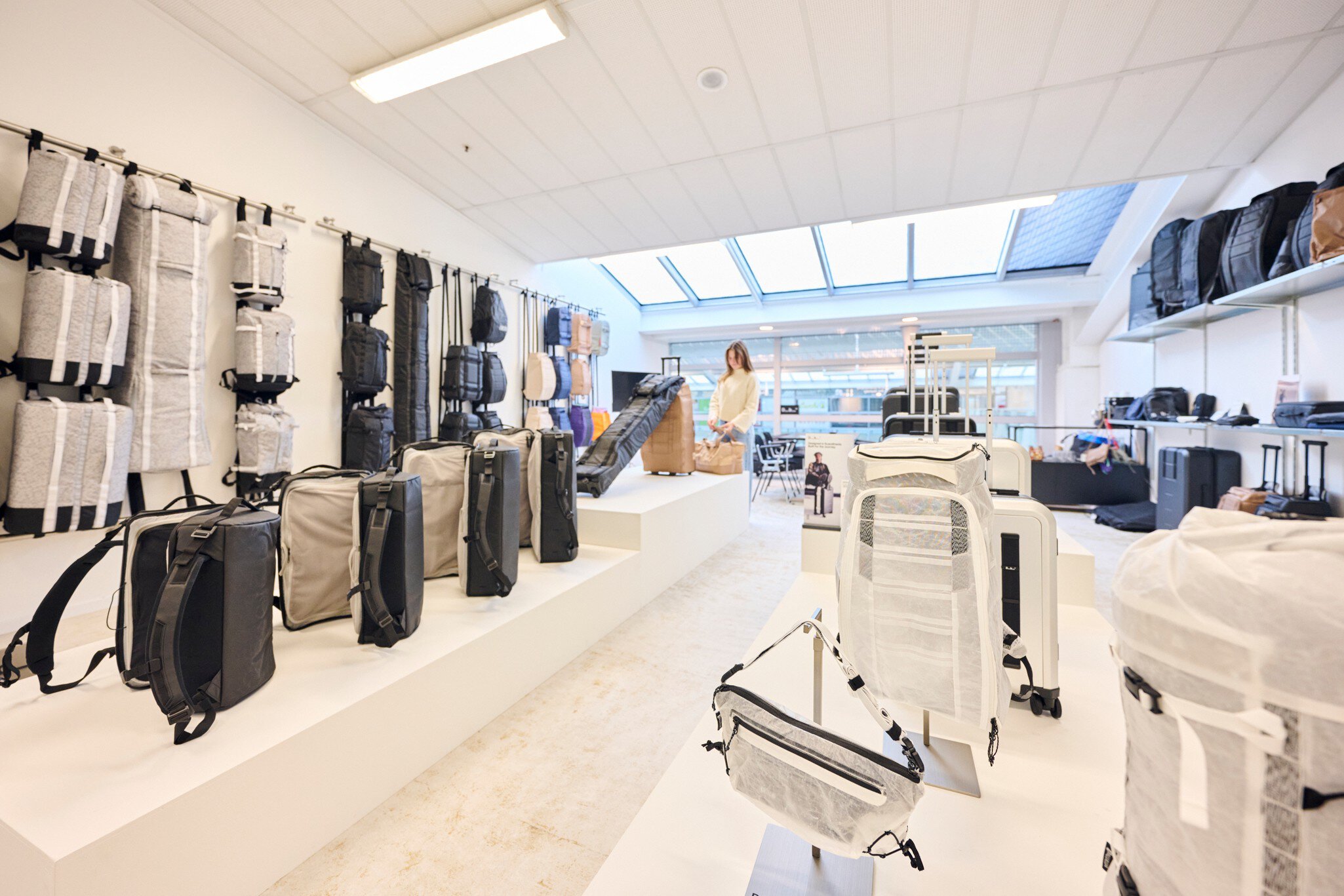 Ein weißer Ausstellungsraum mit hellen Dachfenstern. An den Wänden und auf Tischen werden zahlreiche helle und schwarze Rucksäcke, Taschen und Koffer präsentiert.