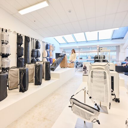 Ein weißer Ausstellungsraum mit hellen Dachfenstern. An den Wänden und auf Tischen werden zahlreiche helle und schwarze Rucksäcke, Taschen und Koffer präsentiert.