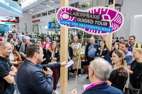Gruppe von Messebesucherinnen und -besuchern, die einem Sprecher lauschen; vorne hält ein Mann ein Schild mit der Aufschrift „LABORPRAXIS GUIDED TOUR – LABOR- UND ANALYSENTECHNIK“.