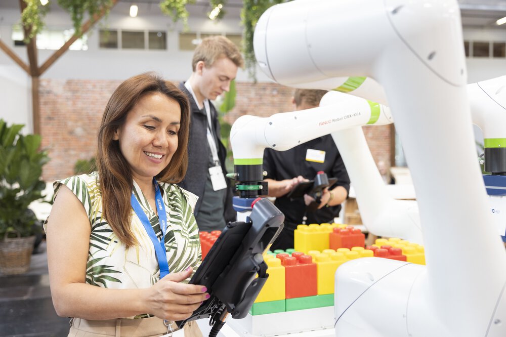 A woman operates a robotic arm at an event; two people and large colorful blocks can be seen in the background.