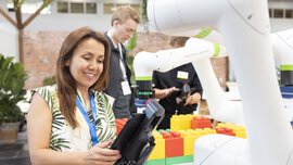 A woman operates a robotic arm at an event; two people and large colorful blocks can be seen in the background.