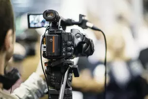 A video camera on a tripod films an event, blurred visitors in the background.