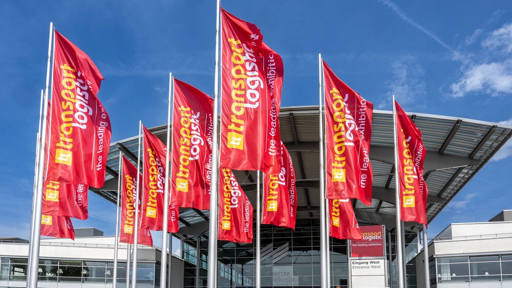 Zahlreiche rote Fahnen der transport logistic wehen vor dem Westeingang zur Messe München. Der Himmel darüber ist blau und leicht bewölkt.