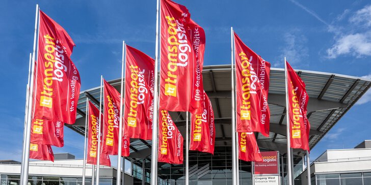 Zahlreiche rote Fahnen der transport logistic wehen vor dem Westeingang zur Messe München. Der Himmel darüber ist blau und leicht bewölkt.