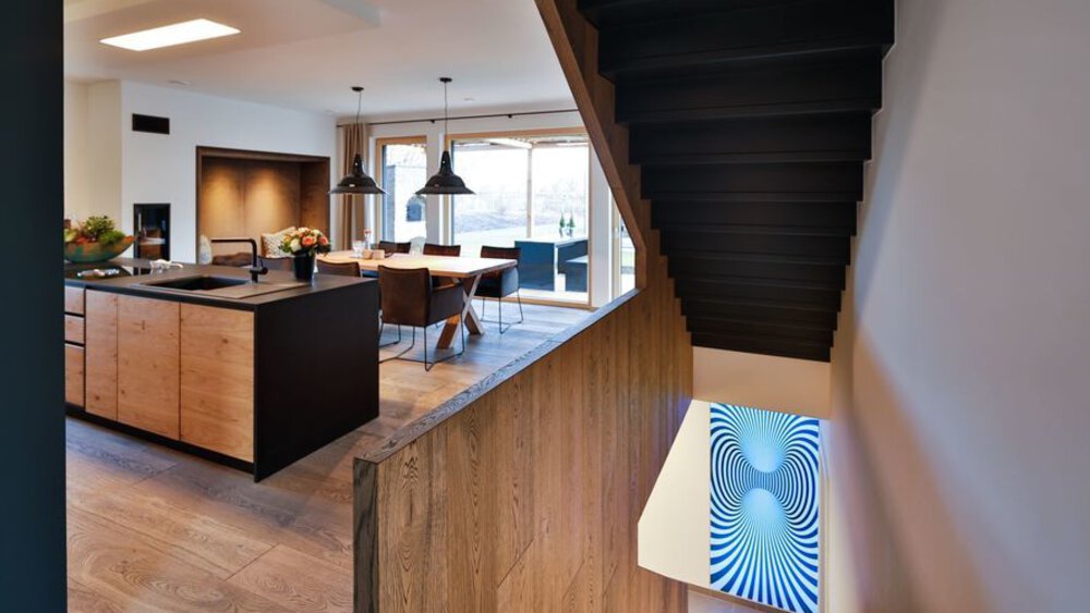 Modernes Interieur mit Kücheninsel, Esstisch und Treppe aus Holz. Abstrakte Kunst an der Wand sorgt für optische Abwechslung.