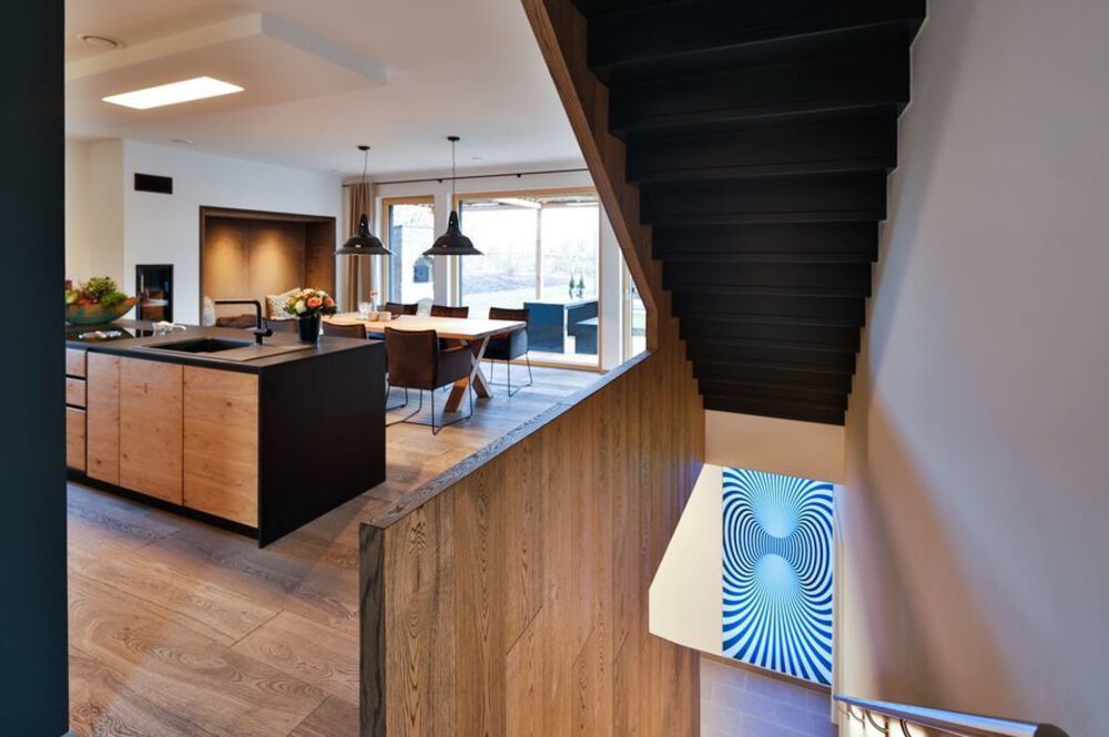 Modernes Interieur mit Kücheninsel, Esstisch und Treppe aus Holz. Abstrakte Kunst an der Wand sorgt für optische Abwechslung.