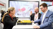 Three trade fair visitors in conversation at a booth, examining an open product case together.