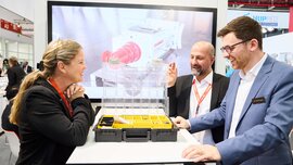 Three trade fair visitors in conversation at a booth, examining an open product case together.