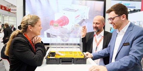 Drei Messebesucher im Gespräch an einem Stand, betrachten gemeinsam eine geöffnete Produktbox.