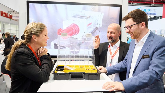 Three trade fair visitors in conversation at a booth, examining an open product case together.