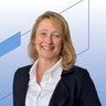 Eine lächelnde Frau mit langen blonden Haaren und hellen Augen trägt eine Halskette, eine weiße Bluse und ein dunkles Jackett.