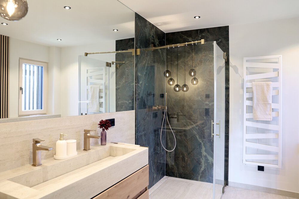 Modernes Badezimmer mit Doppelwaschbecken, Glasdusche mit dunkler Fliesenwand und Handtuch auf einem weißen Handtuchwärmer.