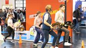 Mehrere Personen, Erwachsene und Kinder, führen Steckenpferde in einem Parcours auf einer Messe; eine Moderatorin mit Headset leitet, Zuschauer im Hintergrund.