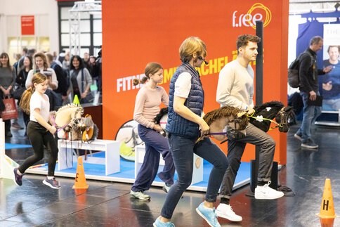 Mehrere Personen, Erwachsene und Kinder, führen Steckenpferde in einem Parcours auf einer Messe; eine Moderatorin mit Headset leitet, Zuschauer im Hintergrund.