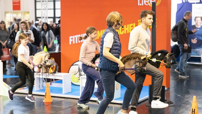 Mehrere Personen, Erwachsene und Kinder, führen Steckenpferde in einem Parcours auf einer Messe; eine Moderatorin mit Headset leitet, Zuschauer im Hintergrund.