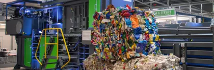 Stacked bales of compressed plastic waste in front of a baler exhibited on a trade fair stand.