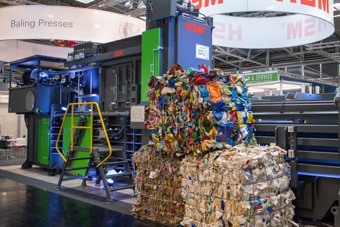 Gestapelte Ballen aus komprimiertem Plastikmüll vor einer, auf einem Messestand ausgestellten Ballenpresse.