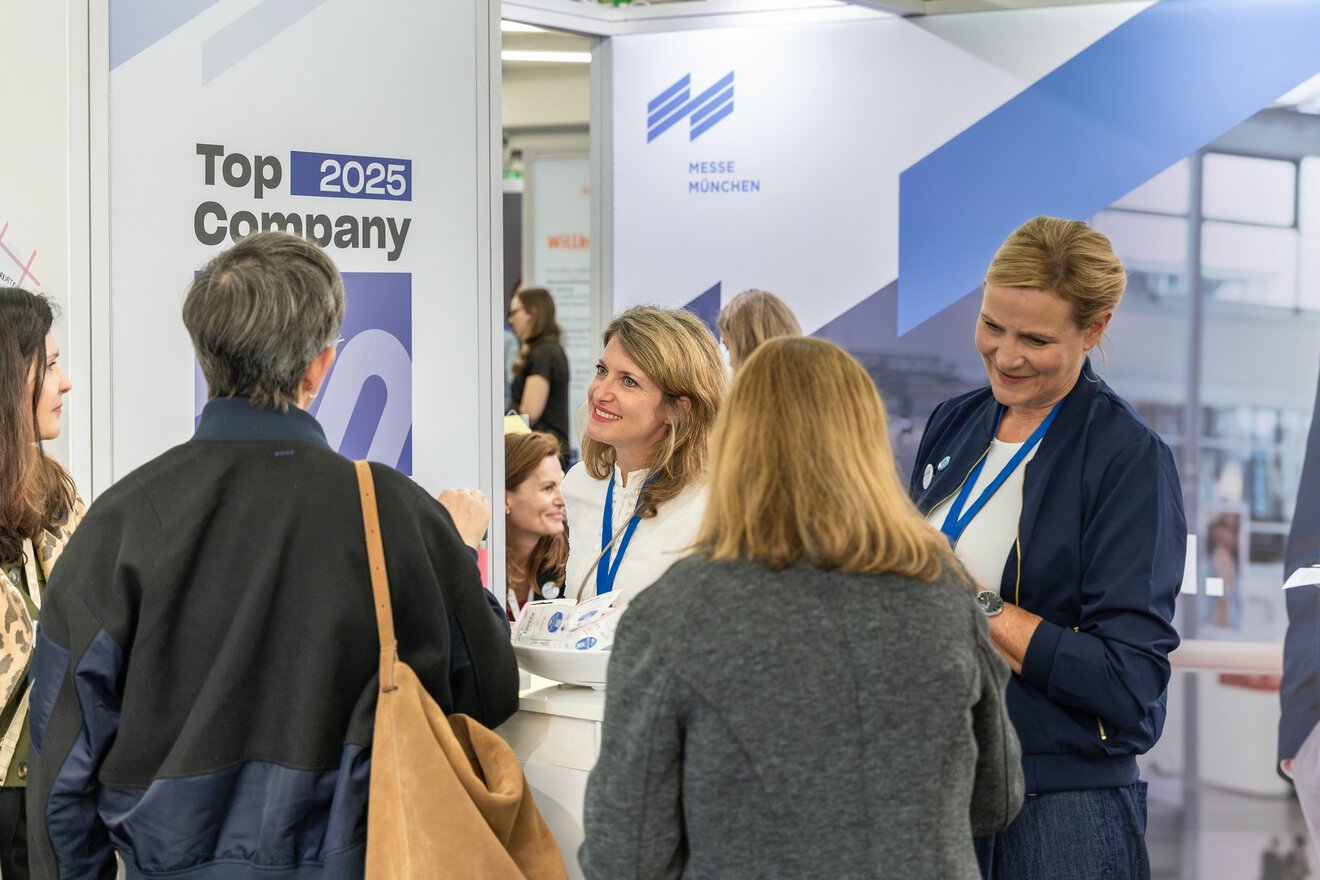 auf dem Bild sieht man Kolleginnen der Messe München auf der Messe herCAREER , sie sind in einem gute beleuchteten Umfeld am Messestand in formeller Kleidung zu sehen.