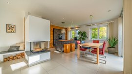 Moderner, offener Wohnbereich mit Kamin, Esstisch, roten Stühlen, Kücheninsel und großen Fenstern, durch die viel Tageslicht hereinströmt.