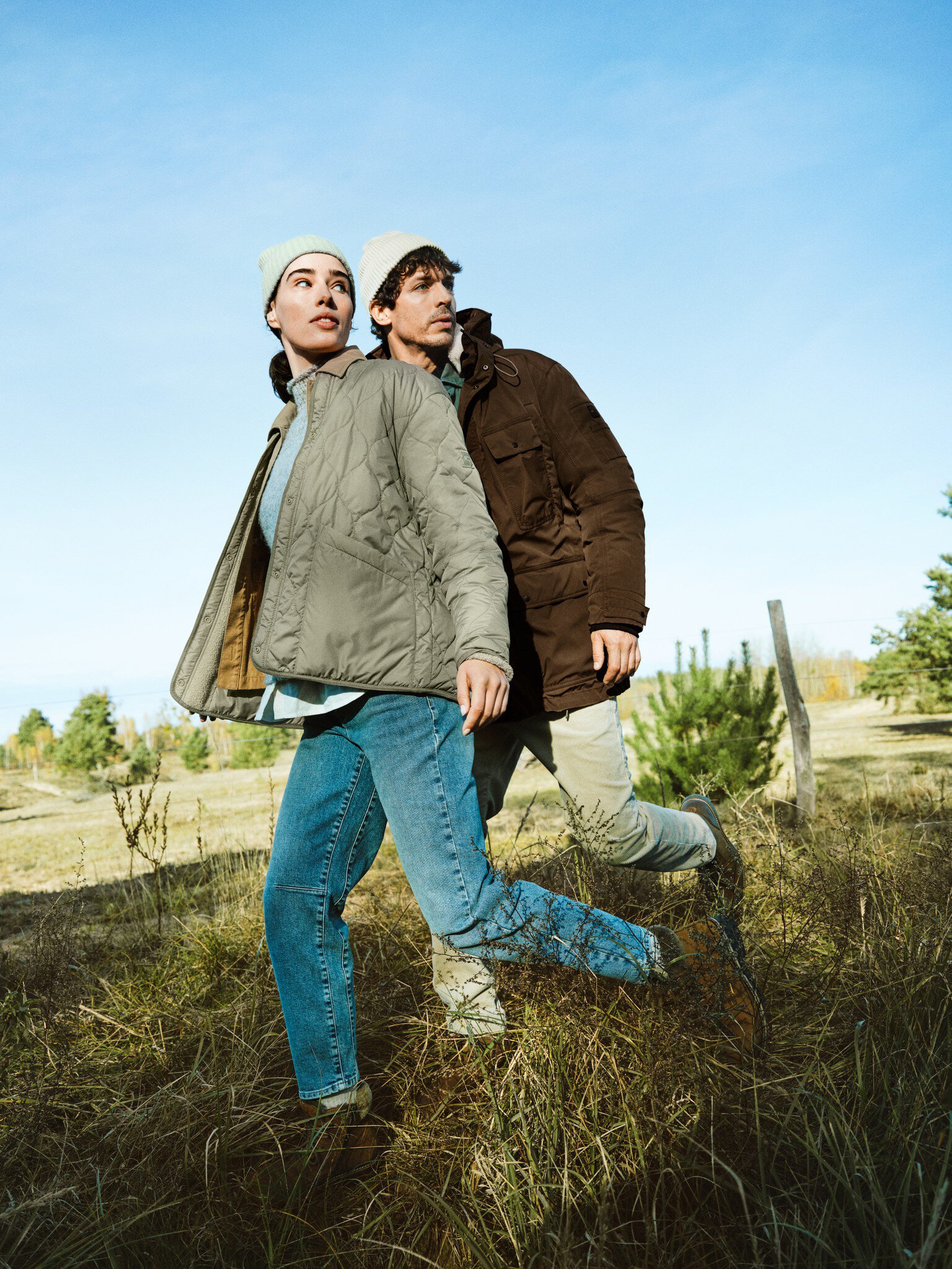 Zwei Personen in Jacken und Mützen laufen an einem sonnigen Tag über eine Wiese und schauen in die Ferne.