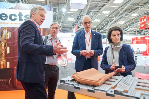 Four trade visitors discuss ceramic products at an exhibition booth while receiving explanations about the displayed components.