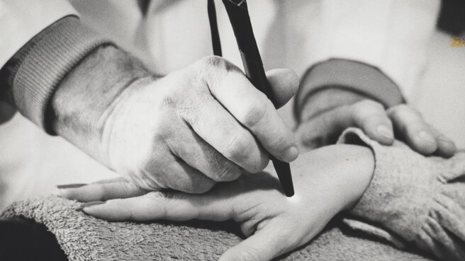 Sharp white photo of a person holding a medical device in the form of a pen to the hand of another person.