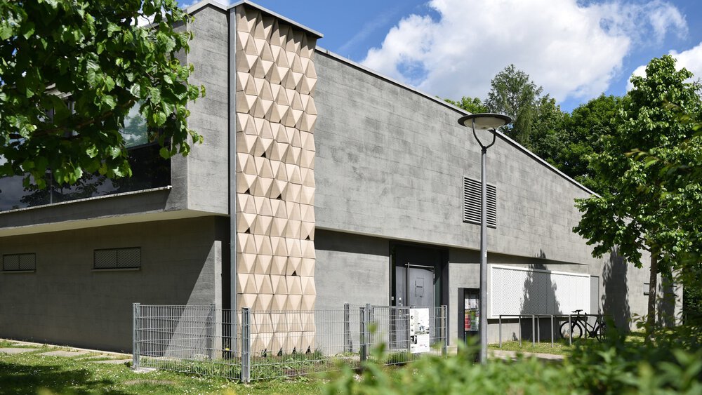 Modernes graues Gebäude mit geometrischer Verkleidung, umgeben von Grün und Bäumen unter einem teilweise bewölkten Himmel.