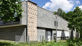 Modernes graues Gebäude mit geometrischer Verkleidung, umgeben von Grün und Bäumen unter einem teilweise bewölkten Himmel.