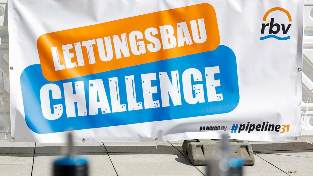 Nahaufnahme eines blauen Rohrleitungsbauteils auf einer Palette, im Hintergrund ein Banner mit der Aufschrift ‚Leitungsbau Challenge.