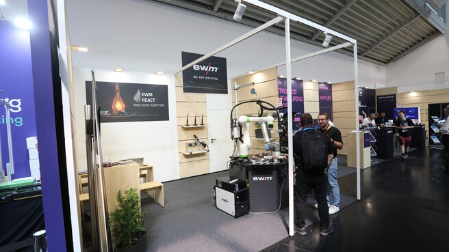 Trade fair booth featuring welding technology, an industrial robot and product displays, with visitors in conversation in an exhibition hall.