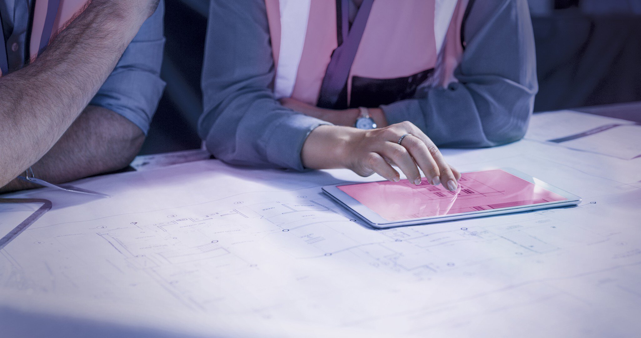 Die Person in der Sicherheitsweste zeigt auf ein Tablet, das auf Architekturplänen liegt.